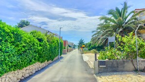 Calle estrecha flanqueada por muros de piedra y abundante vegetación, que conduce a un edificio con tejado rojo.