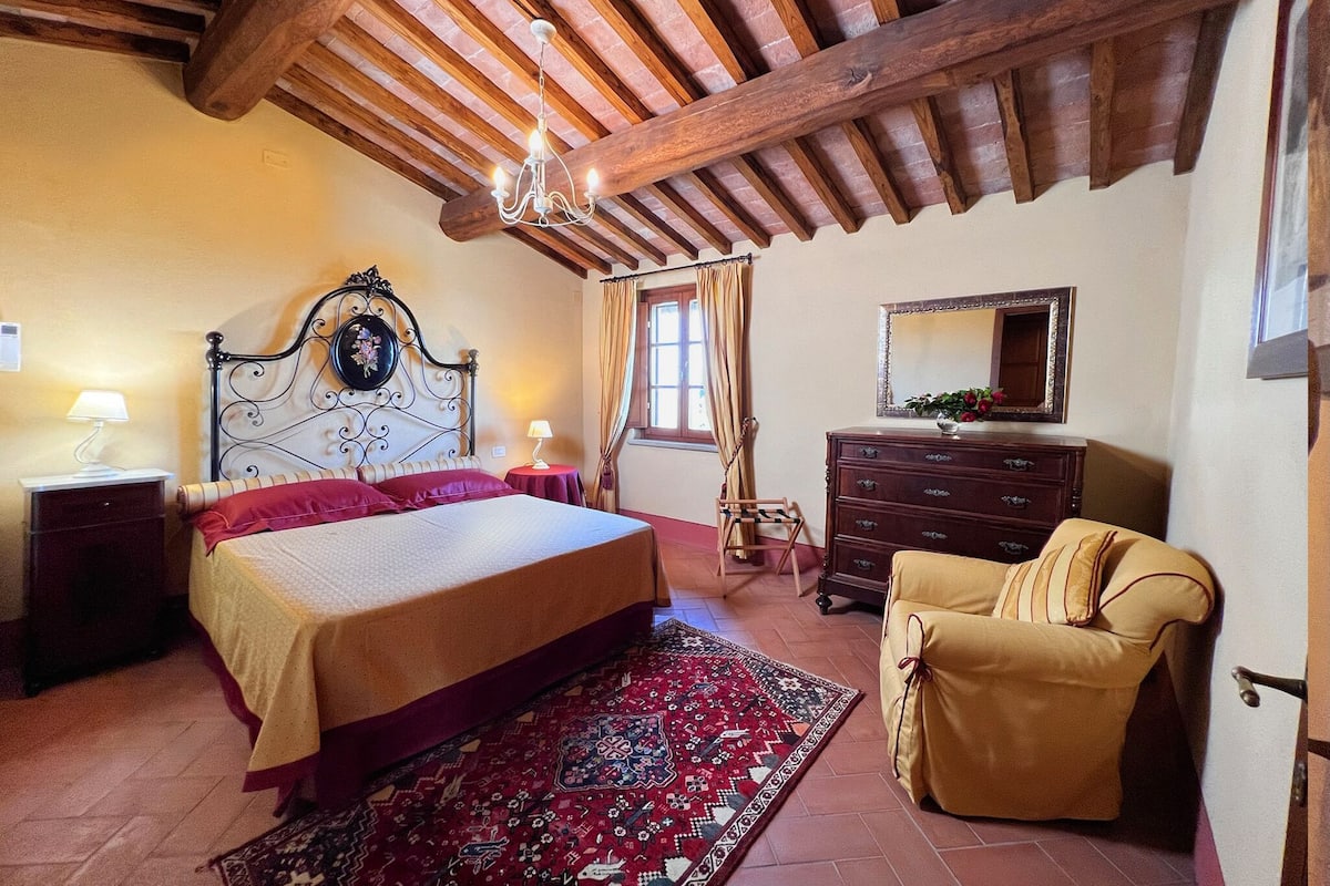 Ein Schlafzimmer mit einem großen Bett, Holzbalkendecke, einem Schminktisch, einem Sessel und einem Spiegel.