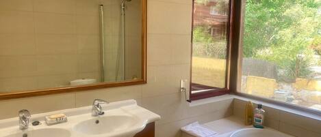 A bathroom with a double sink, a mirror, a window with a view of greenery, and a bathtub.