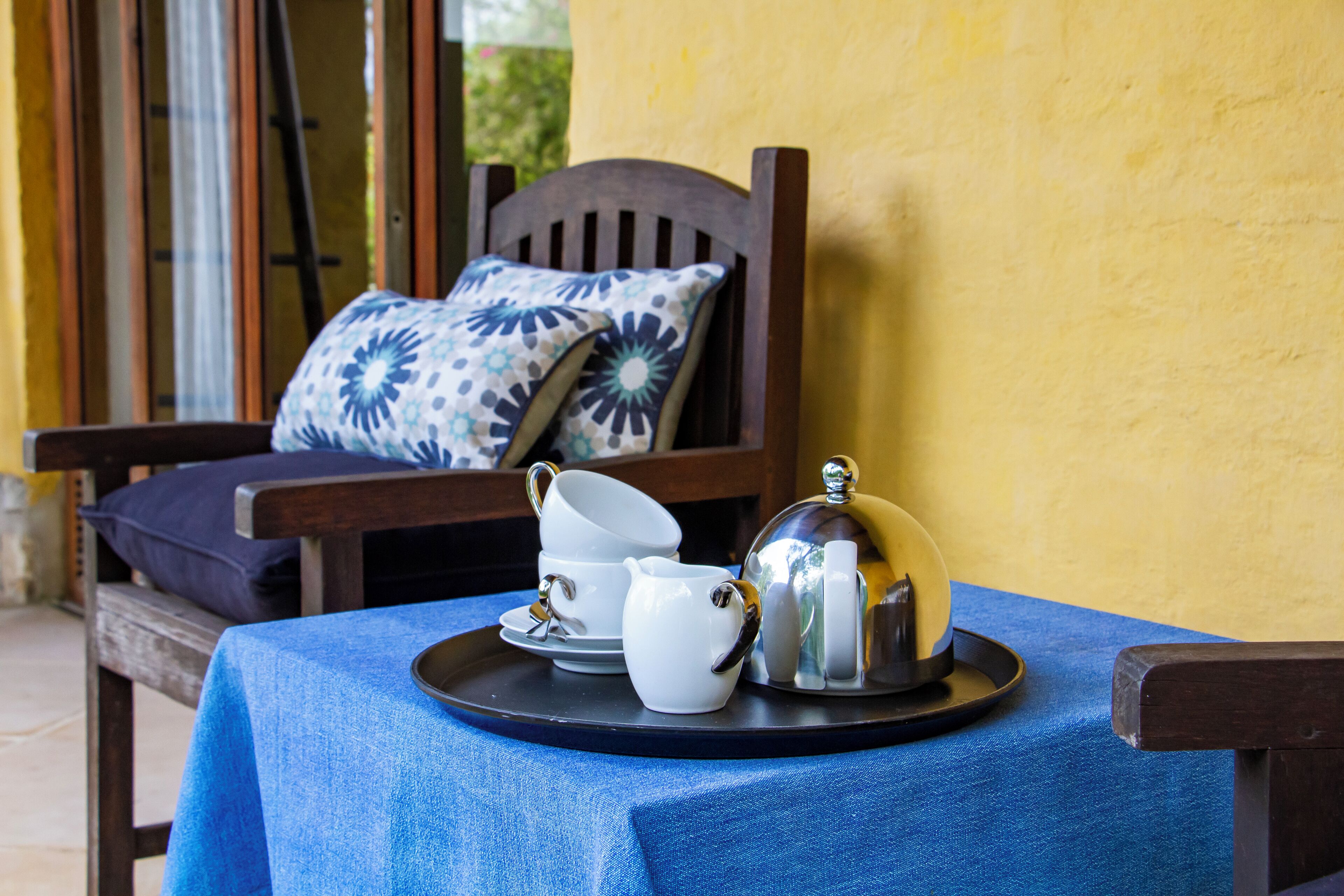 A table with a blue tablecloth, a tray with a teapot, cups, and saucers, and a wooden chair with a patterned pillow.