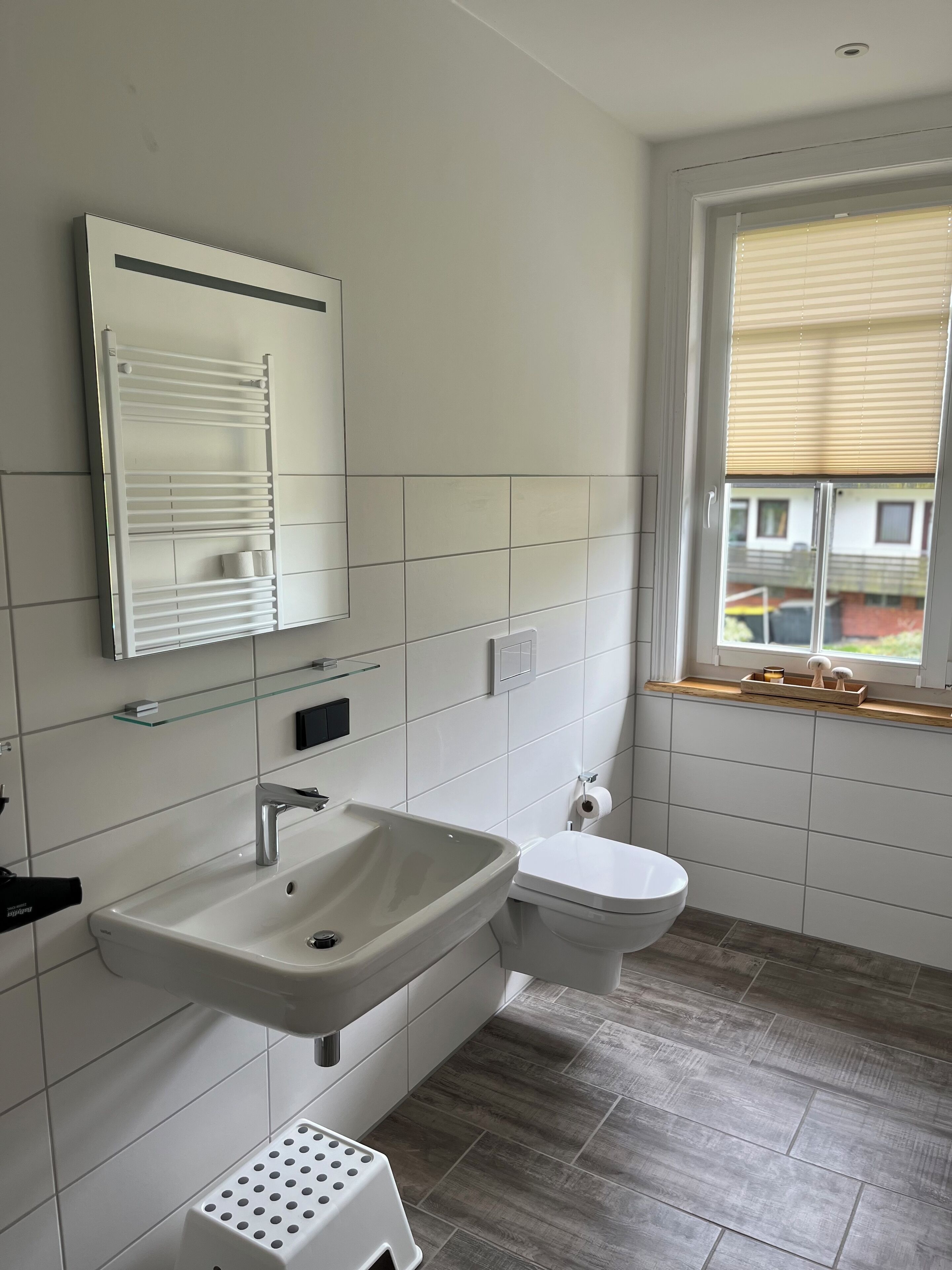 Et moderne badeværelse med hvid vask, spejl og toilet. Der er et vindue med persienner, en lille hylde og trægulv.