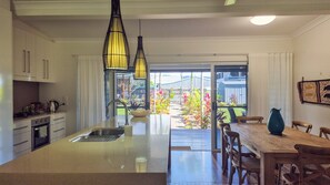 Una cocina con una isla central, un comedor con mesa y sillas de madera, y vistas de un jardín a través de la puerta abierta.