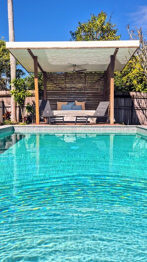 Una piscina cristalina con una zona de estar cubierta y sillones de descanso.