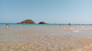 Ein Strand mit klarem Wasser, Felsen und Menschen, die schwimmen und spazieren gehen.