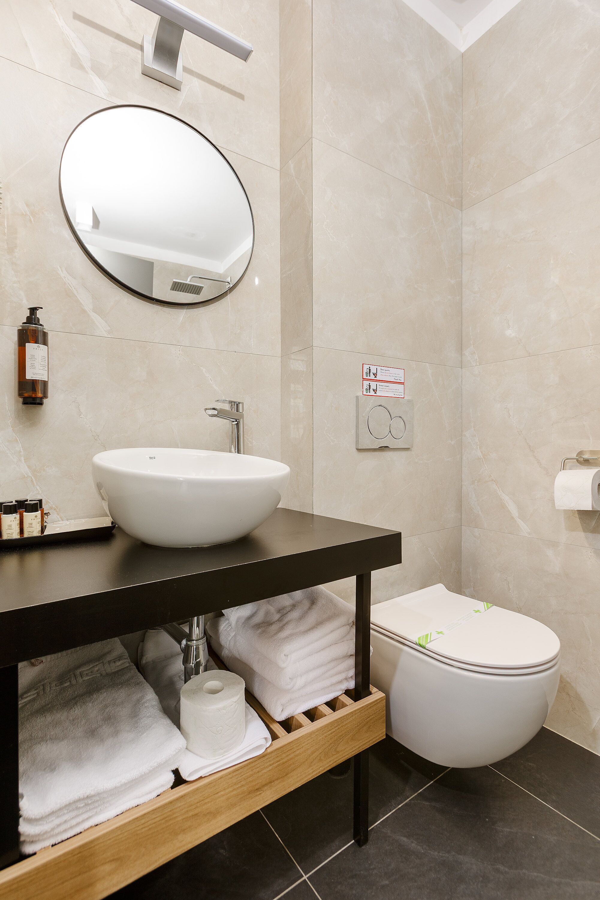 A modern bathroom with a round mirror, a white sink, and a toilet. There are towels and toiletries on the counter.