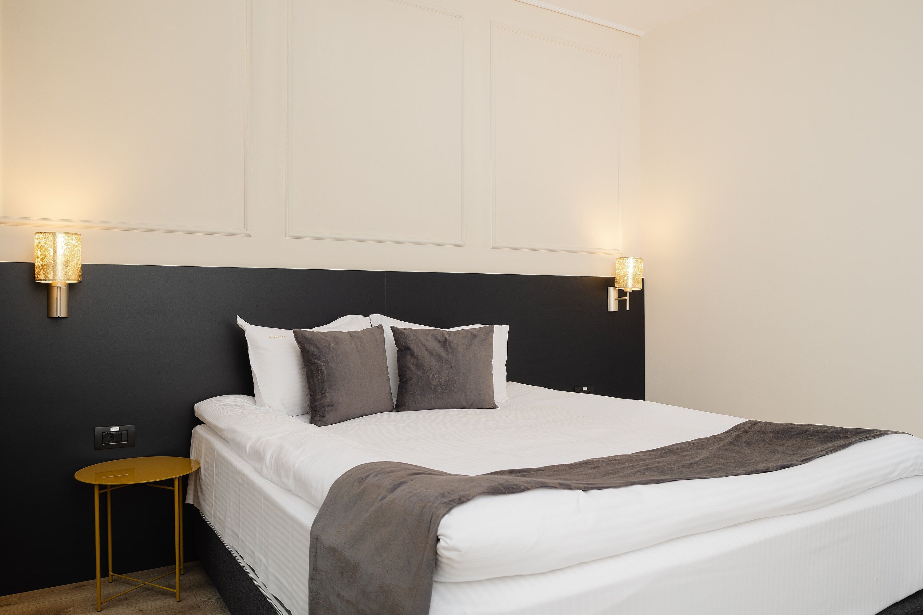 A neatly made bed with a white duvet and a gray throw blanket, flanked by two wall-mounted lamps with a gold finish.