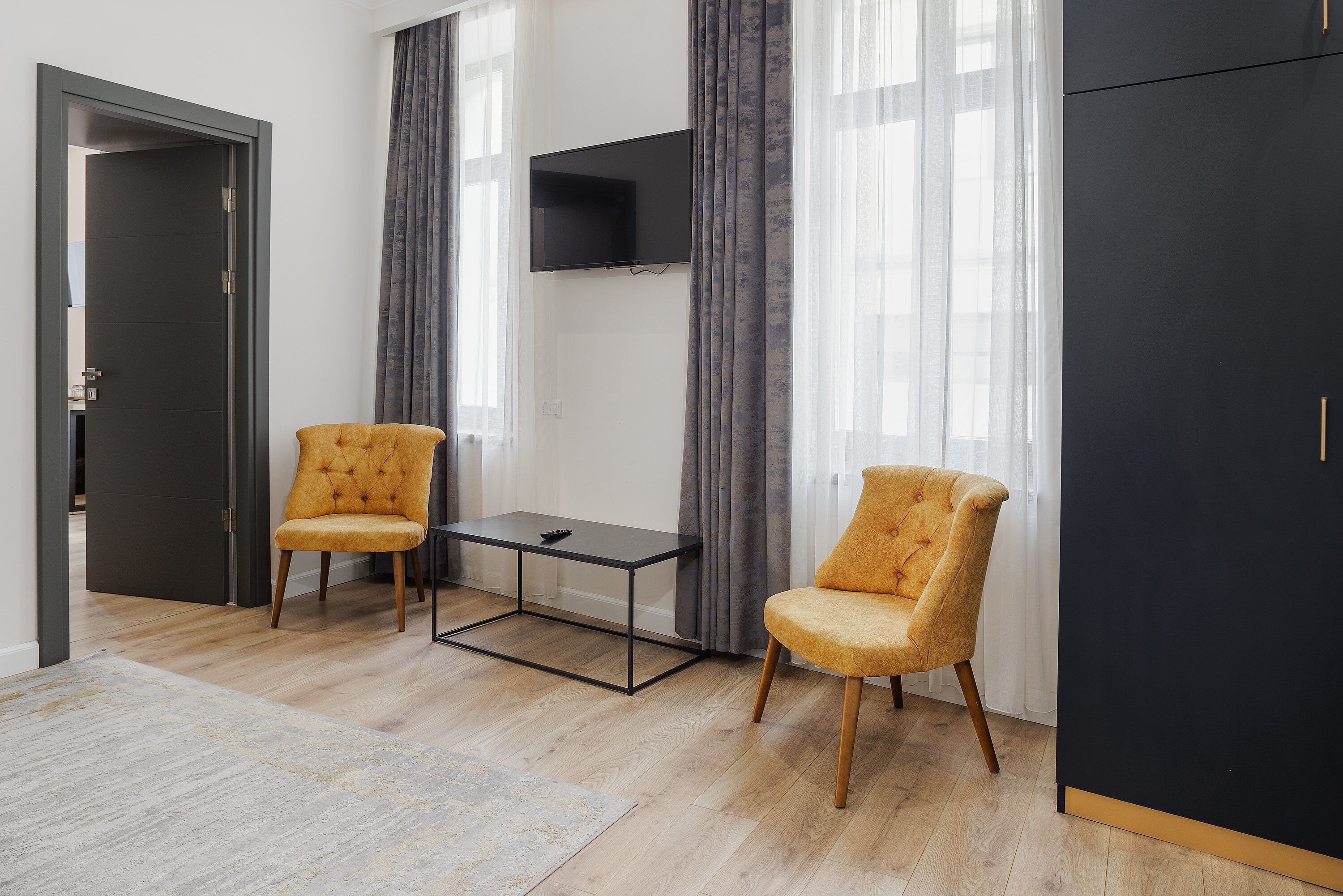Une chambre moderne avec deux fauteuils jaunes, une petite table noire, un téléviseur fixé au mur et une armoire.