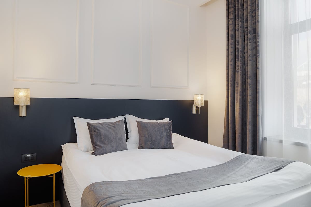 A neatly made bed with white linens and a grey throw, a small yellow side table, and a dark blue headboard.