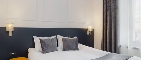 Una cama bien hecha con sábanas blancas y un plaid gris, una mesita de noche amarilla pequeña y un cabecero azul oscuro.