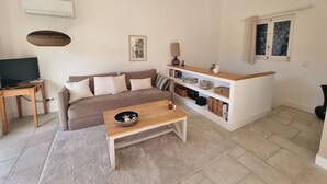 A living room with a brown sofa, a wooden coffee table, a TV on a wooden stand, a bookshelf, and a window with curtains. - Renovated T3 House in Collioure, 2 Bedrooms, Balcony (Collioure)