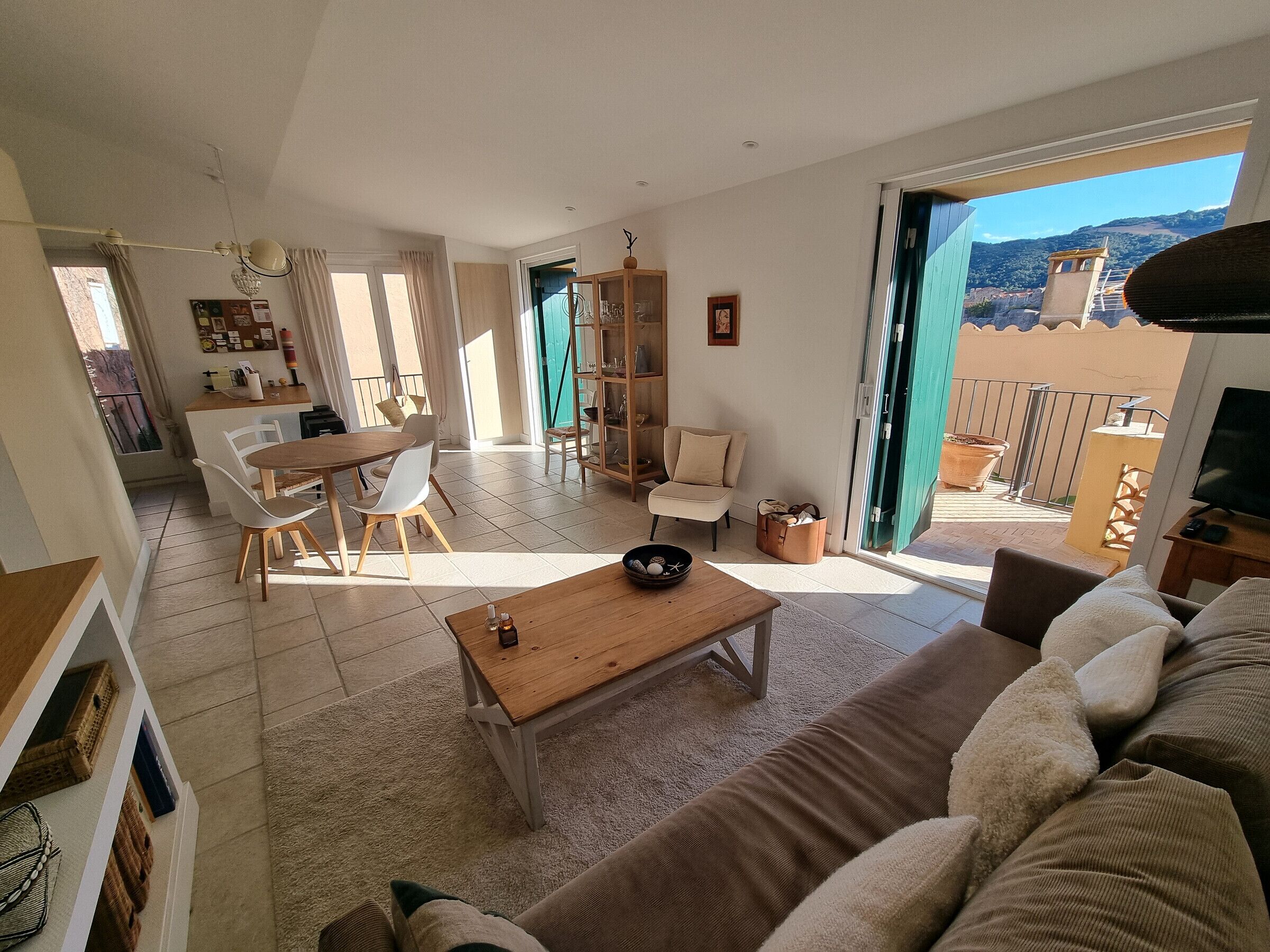 A spacious living area with a sofa, coffee table, and dining set. A sliding glass door leads to a balcony with a view of a mountain and a tower.