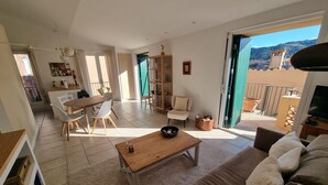 A spacious living area with a sofa, coffee table, and dining set. A sliding glass door leads to a balcony with a view of a mountain and a tower. - Renovated T3 House in Collioure, 2 Bedrooms, Balcony (Collioure)