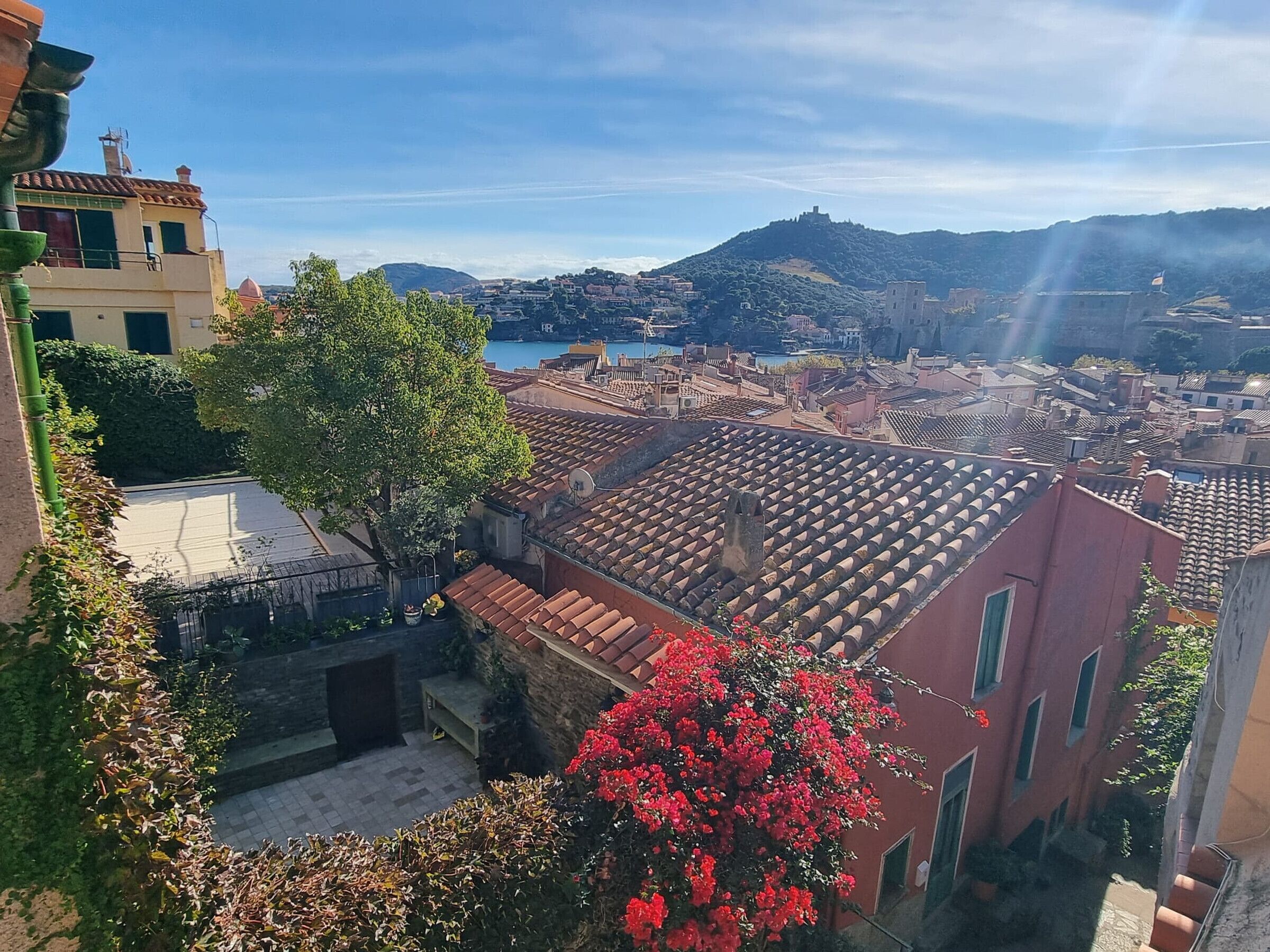 A view of a coastal town with terracotta rooftops, a prominent tree, and a distant hill with a castle-like structure.