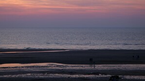 A beach at sunset with calm waters and a few people walking on the sand.