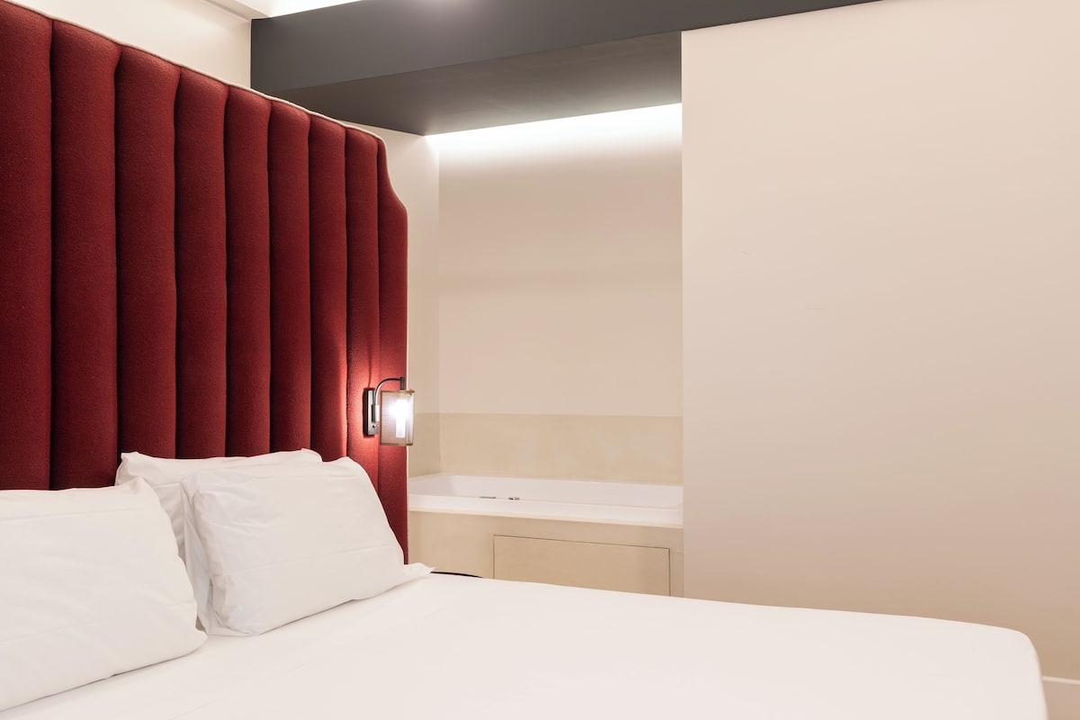 A hotel room with a bed, white pillows, a red headboard, a wall-mounted light, and a small white bedside table.