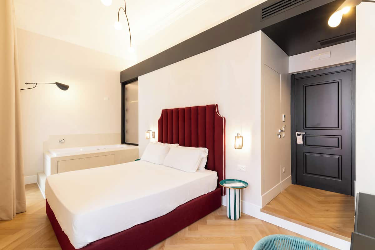 A modern bedroom with a large bed, a red headboard, a small bedside table, and a black door.