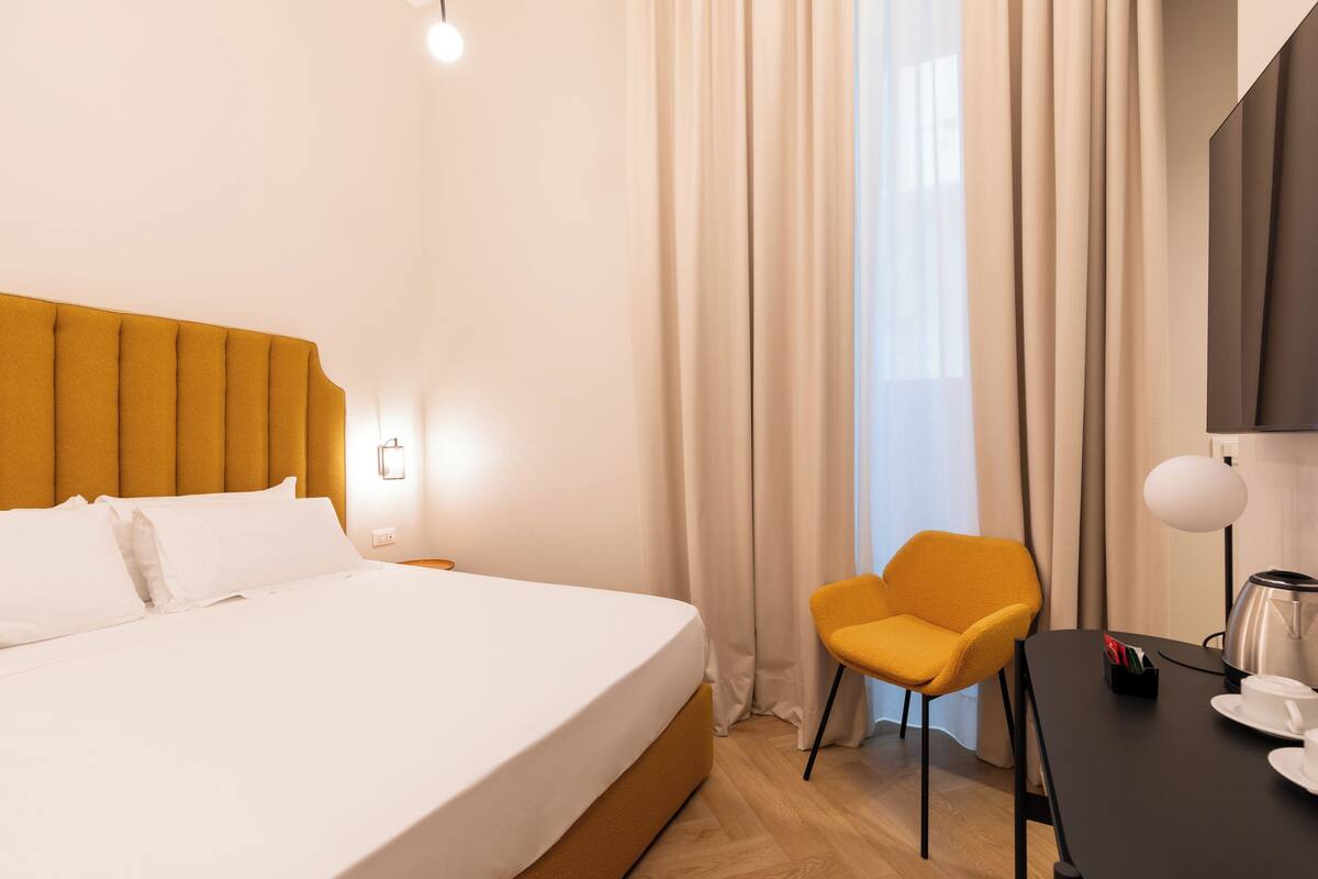 A modern hotel room with a large bed, a yellow armchair, a small table with a kettle, and a large window with curtains.