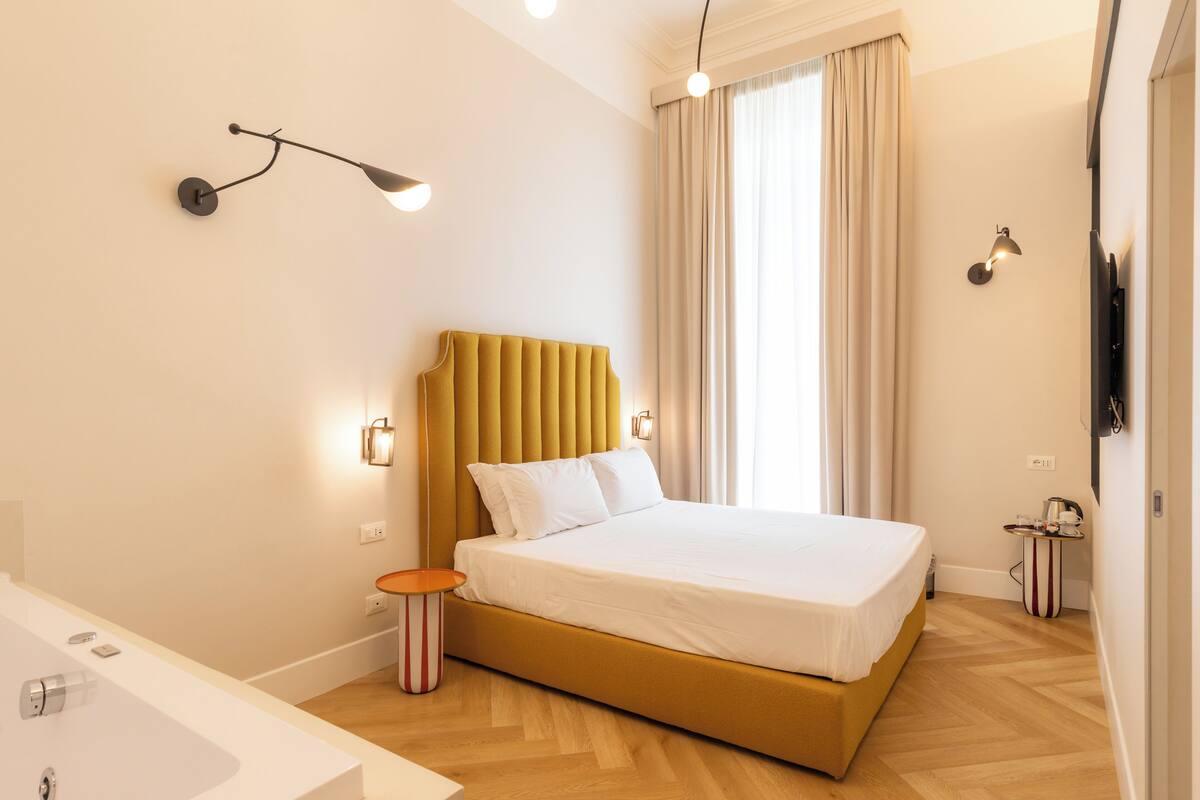 A modern hotel room with a large bed, a bedside table, a wall-mounted TV, and a bathroom with a sink and mirror.