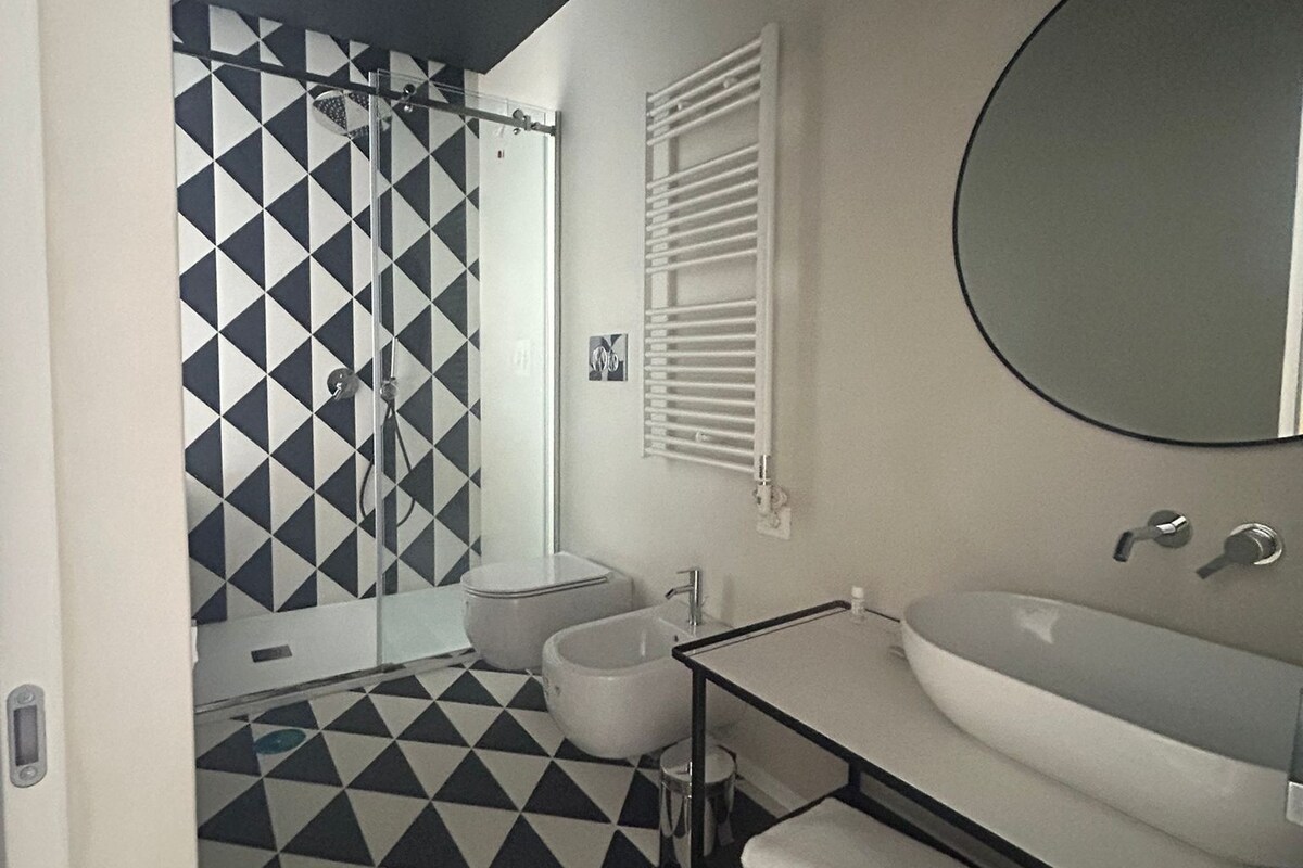 Un baño con piso de baldosas blanco y negro geométricas, un lavamanos blanco con espejo y zona de ducha.