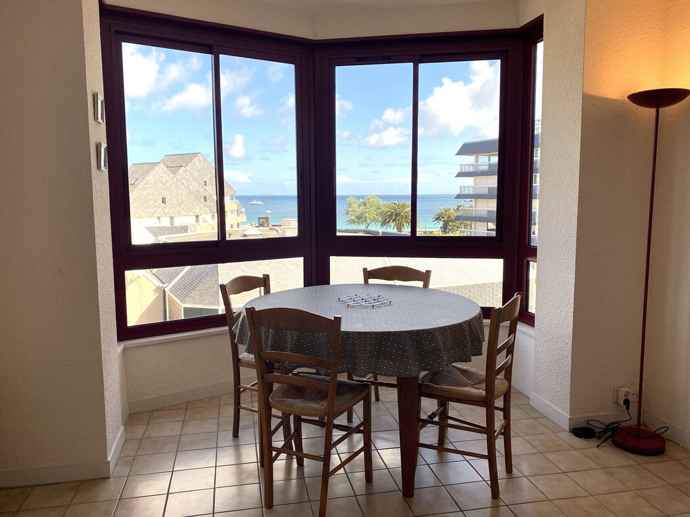 Habitación esquinera con ventanal panorámico que ofrece vistas al mar, una mesa redonda con sillas y una lámpara de pie.