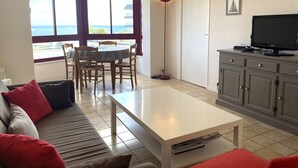 A living room with a sofa, a coffee table, a TV on a cabinet, a dining area with chairs and a table, and a large window with a view of the sea.