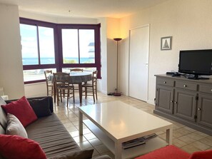 Uma sala de estar com sofá, mesa de centro, televisão sobre um móvel, área de jantar com cadeiras e mesa, e uma grande janela com vista para o mar.
