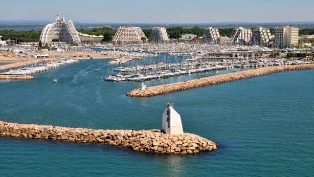 A coastal marina with multiple boats docked, a rocky breakwater, and modern buildings in the background. - Stylish 1-Bed Apt in La Grande-Motte: Modern, Spacious, Loggia, Parking, Near Beach & Shops (La Grande-Motte)