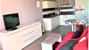 A modern living room with a flat-screen TV, a red sofa, and a kitchen area with white cabinets and a dining table. - Stylish 1-Bed Apt in La Grande-Motte: Modern, Spacious, Loggia, Parking, Near Beach & Shops (La Grande-Motte)