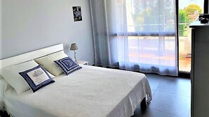 A bedroom with a bed, a nightstand, a lamp, and a window with curtains. - Stylish 1-Bed Apt in La Grande-Motte: Modern, Spacious, Loggia, Parking, Near Beach & Shops (La Grande-Motte)