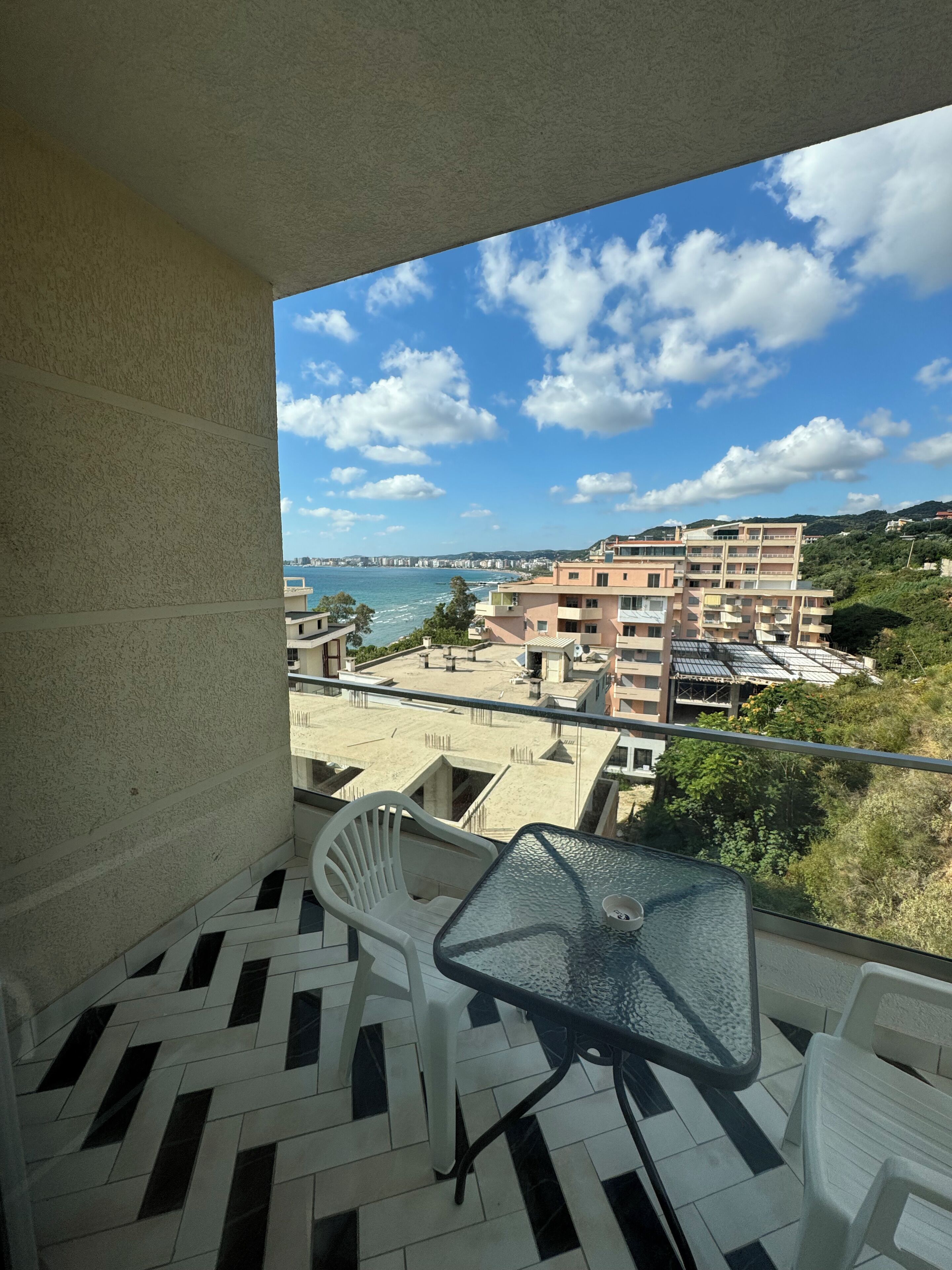 A balcony with a table and chairs, overlooking a coastal cityscape with buildings and a clear sky.
