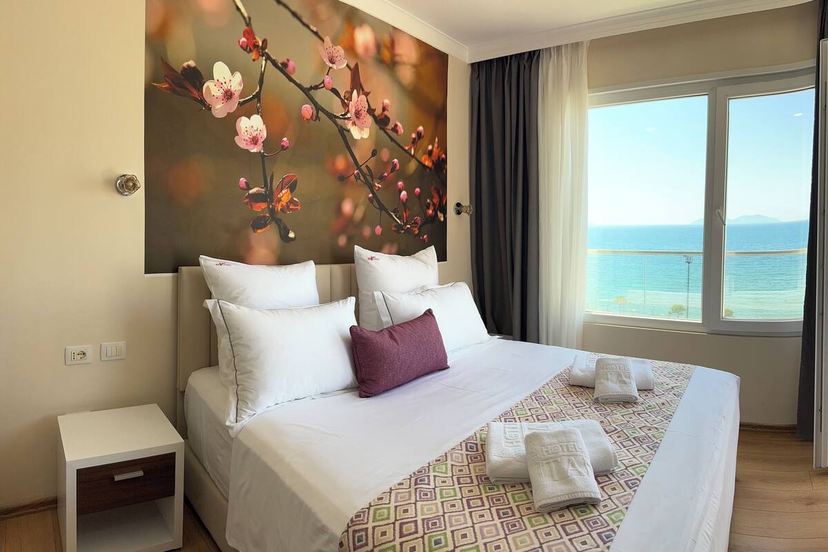 Quarto de hotel com uma cama grande, uma mesa de cabeceira, um quadro de flores na parede, uma janela com vista para o mar e um tapete estampado.