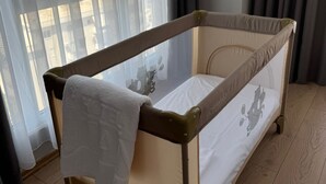 A baby crib with a beige frame and white mattress, positioned near a window with sheer curtains. - Hotel Britania  (Vlorë)