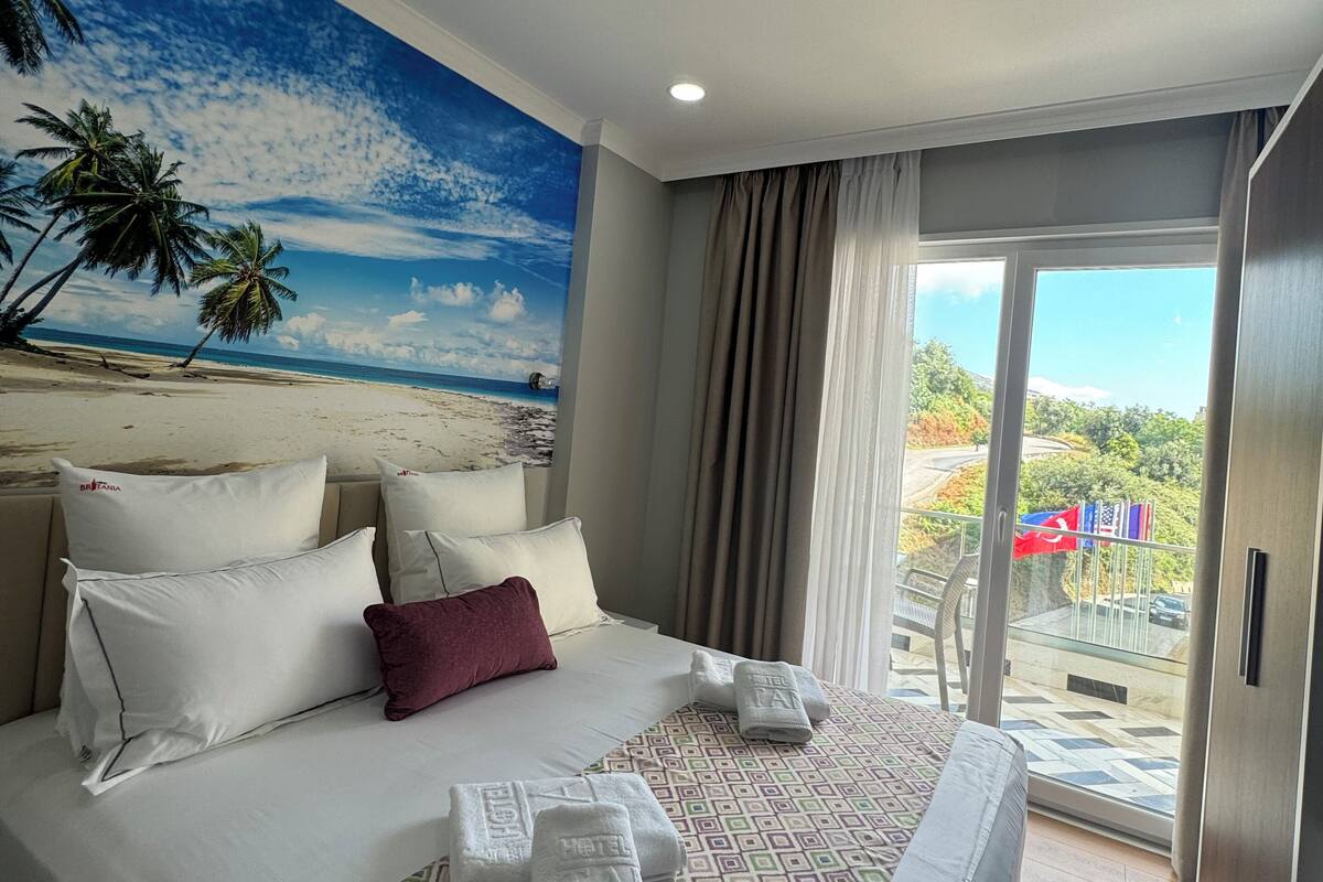 Quarto de hotel com uma cama, uma janela grande e um mural de praia na parede.