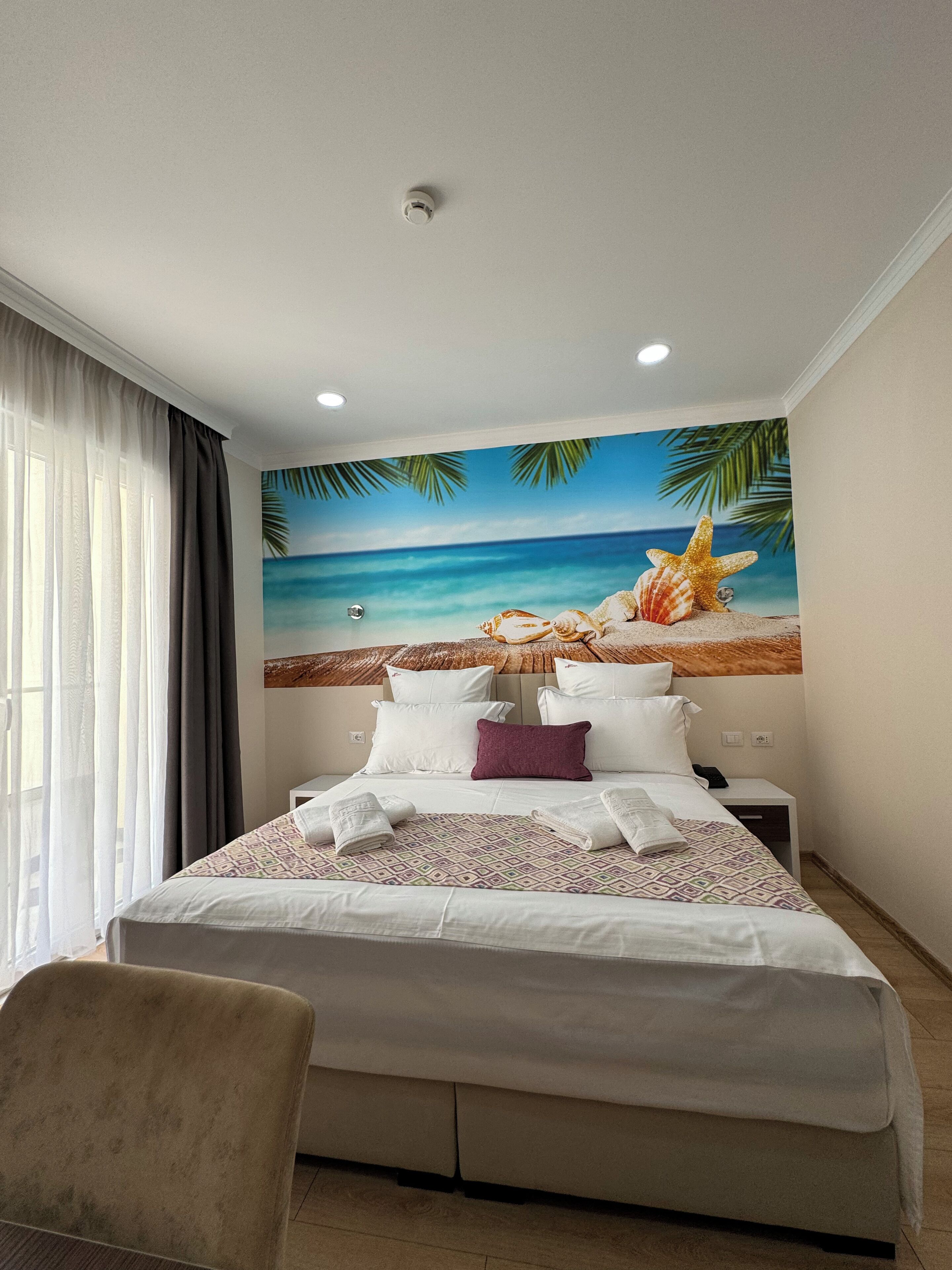 A hotel room with a bed, a chair, a nightstand, and a wall mural of a beach scene.