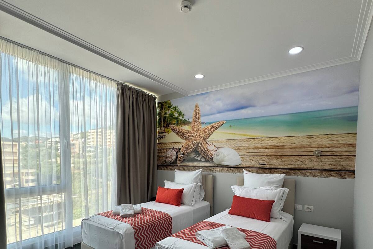 Quarto de hotel com duas camas, uma janela grande com cortinas, uma decoração de estrela-do-mar e um mural de praia na parede.