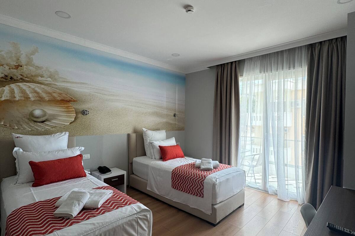 Quarto de hotel com duas camas, um mural com cena de praia, uma escrivaninha e uma janela grande com cortinas.