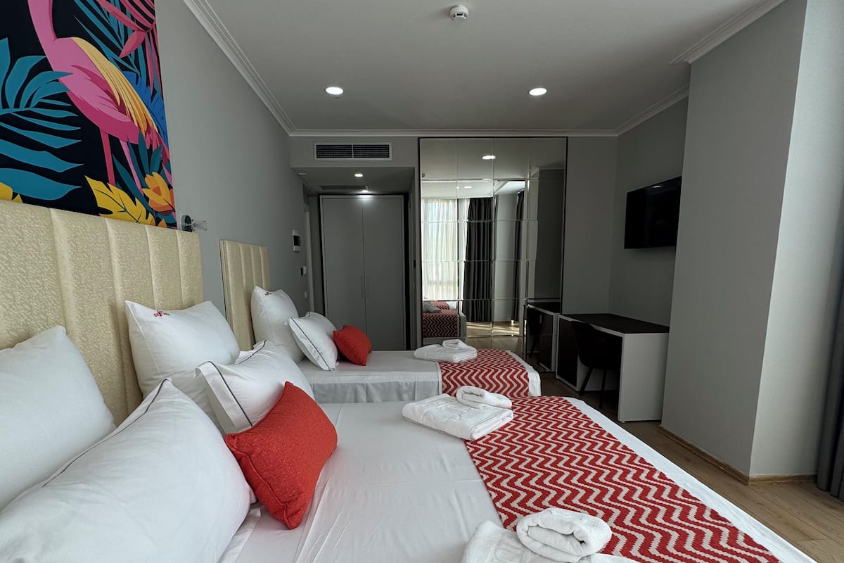 Quarto de hotel com duas camas, um espelho grande, uma televisão e um tapete vermelho e branco com padrão.