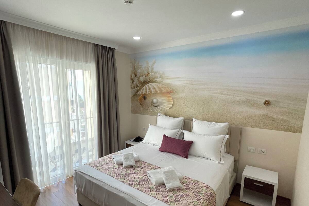 Quarto de hotel com uma cama grande, uma escrivaninha com cadeira, mesa de cabeceira e um mural na parede com uma paisagem praiana.