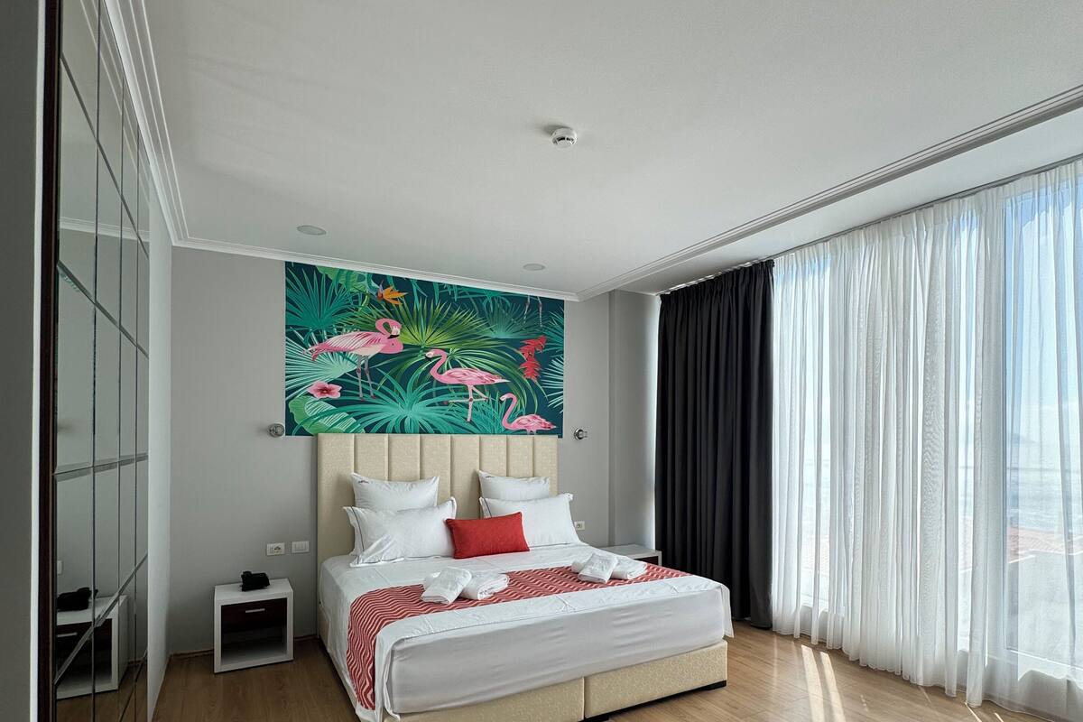 Quarto de hotel moderno com uma cama grande, uma mesa de cabeceira, uma pequena mesa, um espelho e uma janela ampla com cortinas translúcidas.
