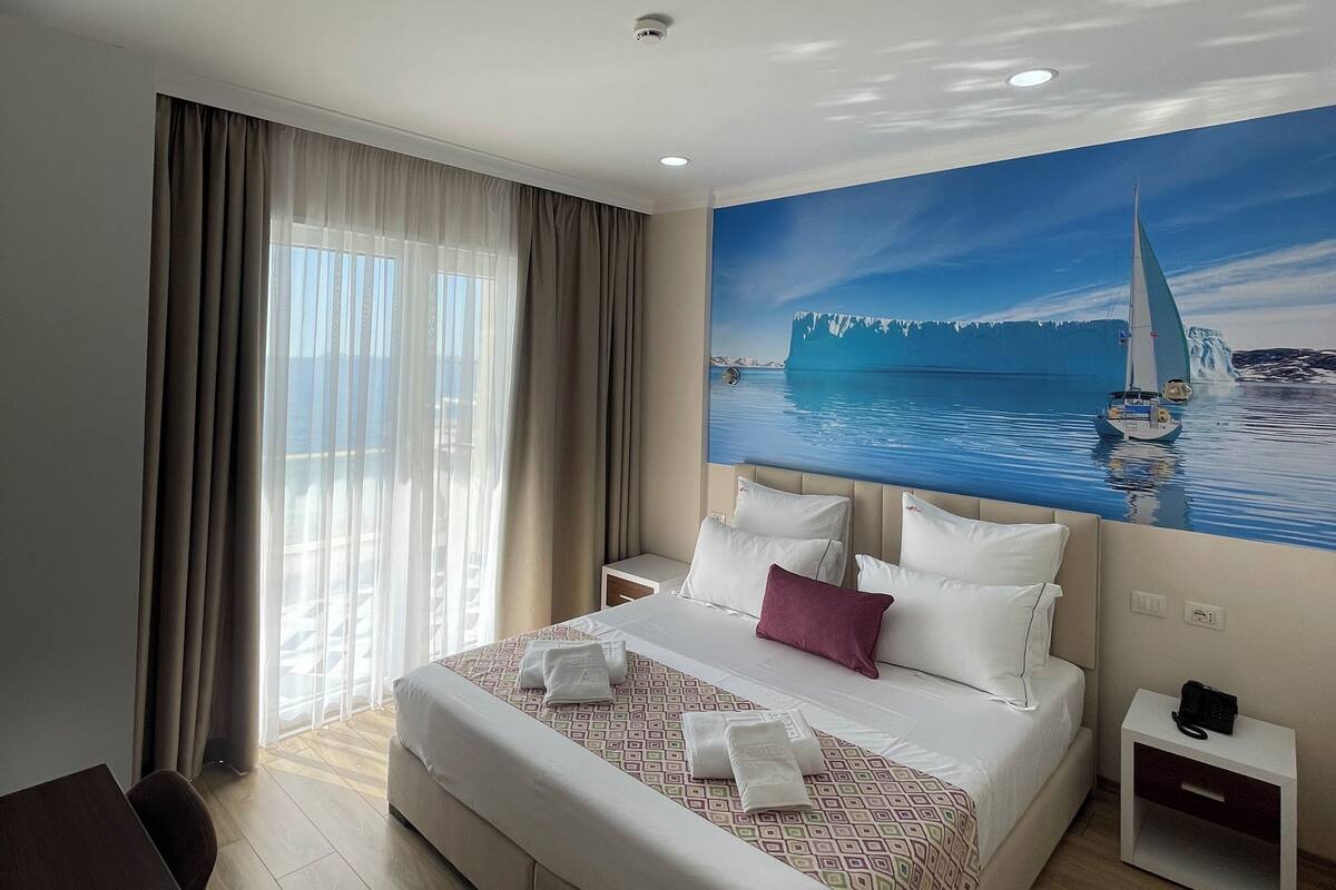Quarto de hotel com uma cama grande, um quadro de um veleiro na parede, uma escrivaninha com um controle remoto e uma mesinha de cabeceira com um telefone.