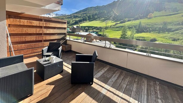 Un balcon doté de meubles en osier et d’un mur en bois, offrant une vue sur une vallée bordée de montagnes et surplombant un ciel dégagé.