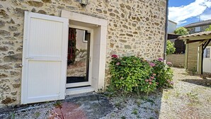 A stone building with a white door open, revealing a garden with green foliage and pink flowers. - Charming F4 House, Terrace, 650m from Lamalou-les-Bains Thermes, Pet-Friendly (Lamalou-les-Bains)