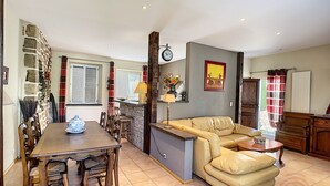 A dining area with a table and chairs, a sofa, a clock, and a kitchen area with a stove. - Charming F4 House, Terrace, 650m from Lamalou-les-Bains Thermes, Pet-Friendly (Lamalou-les-Bains)