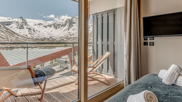 Hotelkamer met groot raam en uitzicht op de bergen, een houten terras buiten, en een bed met een blauwe deken en witte handdoeken.