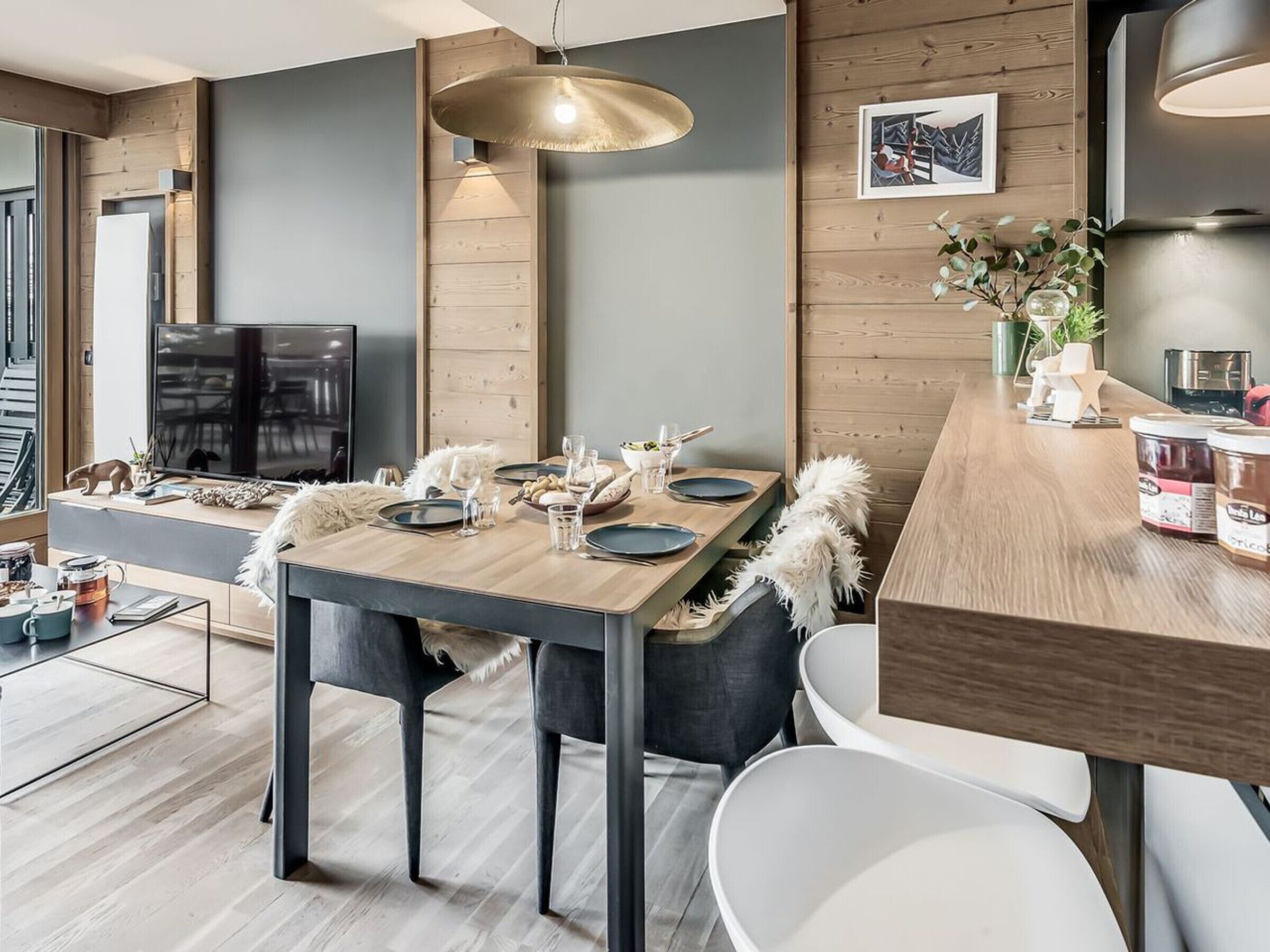 Un espace repas moderne avec une table en bois dressée pour quatre personnes, un téléviseur en arrière-plan et un mur en bois orné d’un cadre.