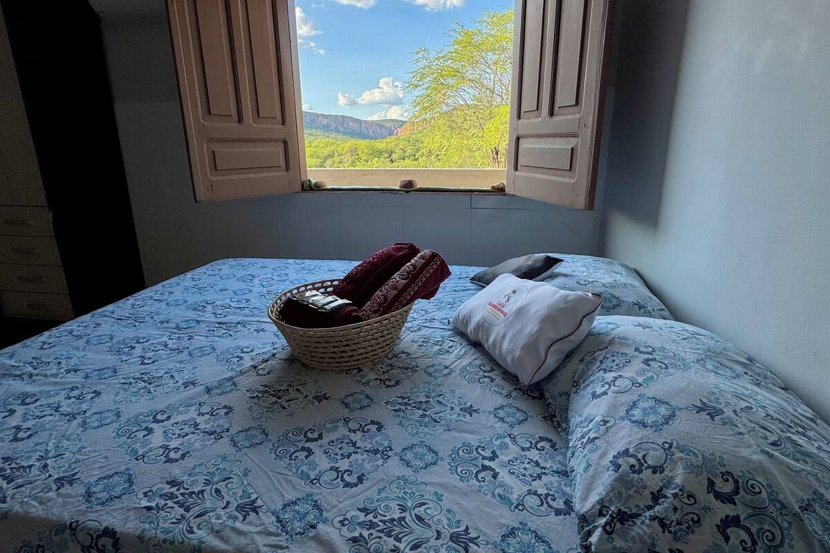 Een slaapkamer met een bed, een mand met handdoeken en een raam met uitzicht op groen en bergen.