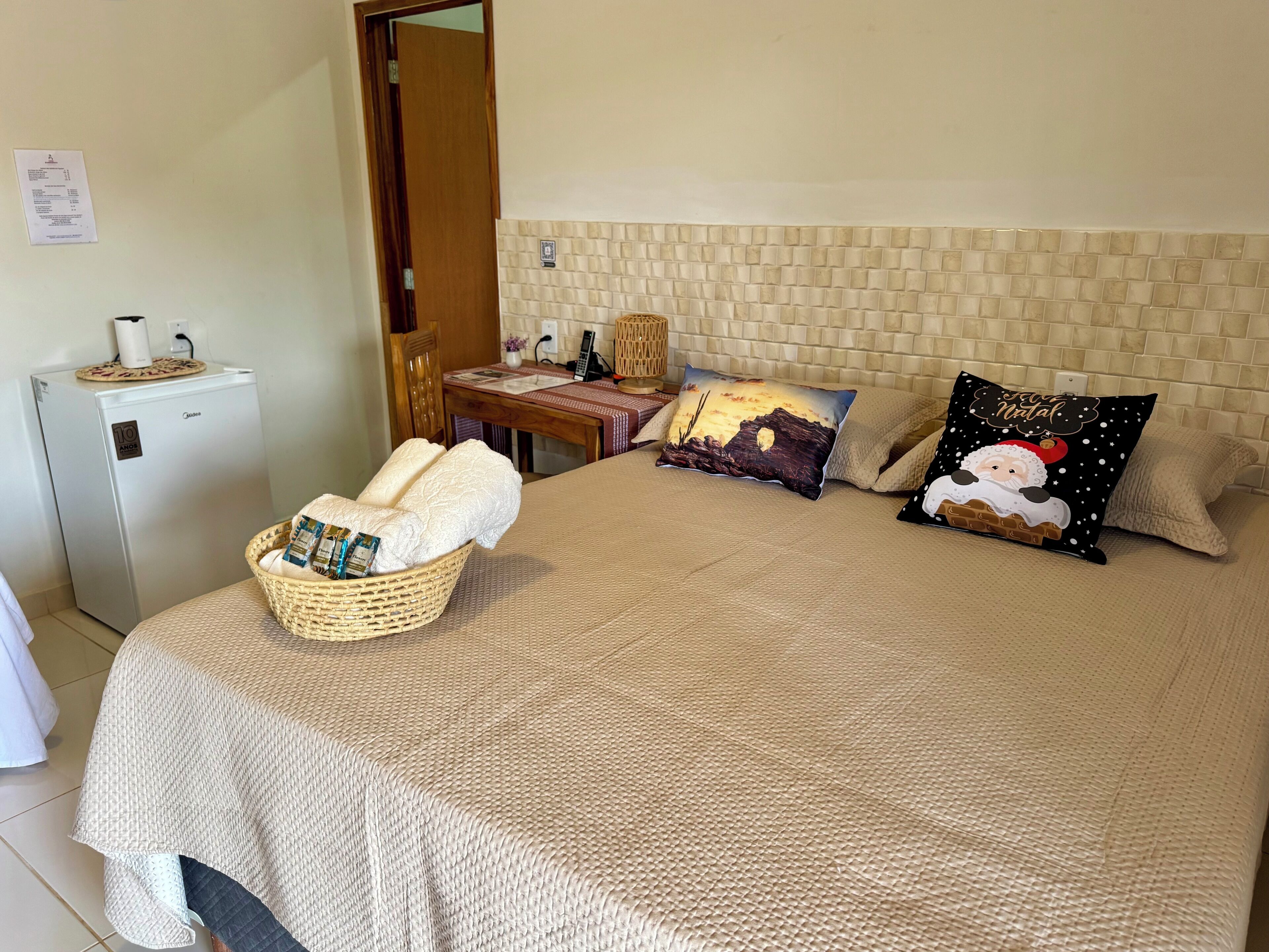Un dormitorio ordenado con cama, almohadas, una mesita, una silla, una cesta y un refrigerador.