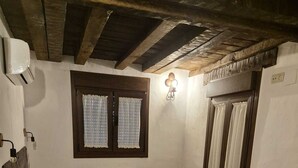 A small, cozy room with a single bed, a radiator, a wooden headboard, a window with curtains, and a wooden stool.