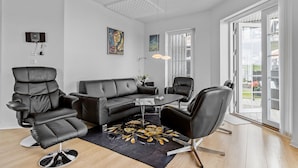 Moderni olohuone, jossa on musta nahkasohva, sohvapöytä ja musta, kukkakuviollinen matto. Huoneessa on kaksi mustaa nahkaista nojatuolia, lattiavalaisin ja seinällä taulu. Huoneessa on suuret ikkunat ja lasiliukuovi, joka johtaa ulkotilaan.