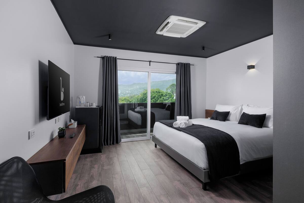 A modern hotel room with a bed, a desk, a chair, a TV, and a balcony with a view of the mountains.